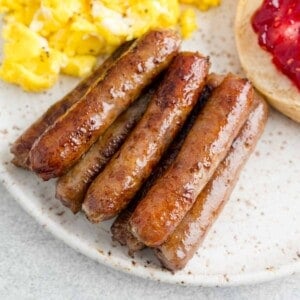 Close-up of a plate with breakfast sausages, scrambled eggs, and a bagel half topped with strawberry jam.