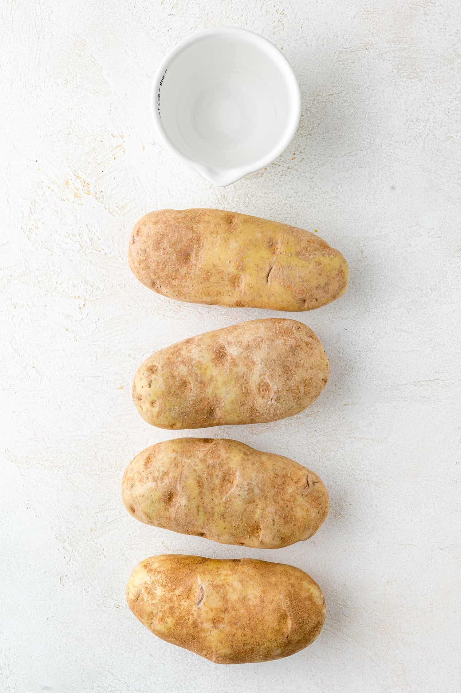 Four potatoes lined vertically on a light surface with a small white bowl above them.