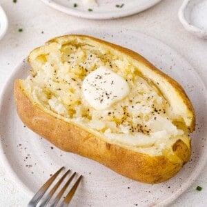 Baked potato with a dollop of butter and sprinkled with black pepper on a white plate, a fork nearby.