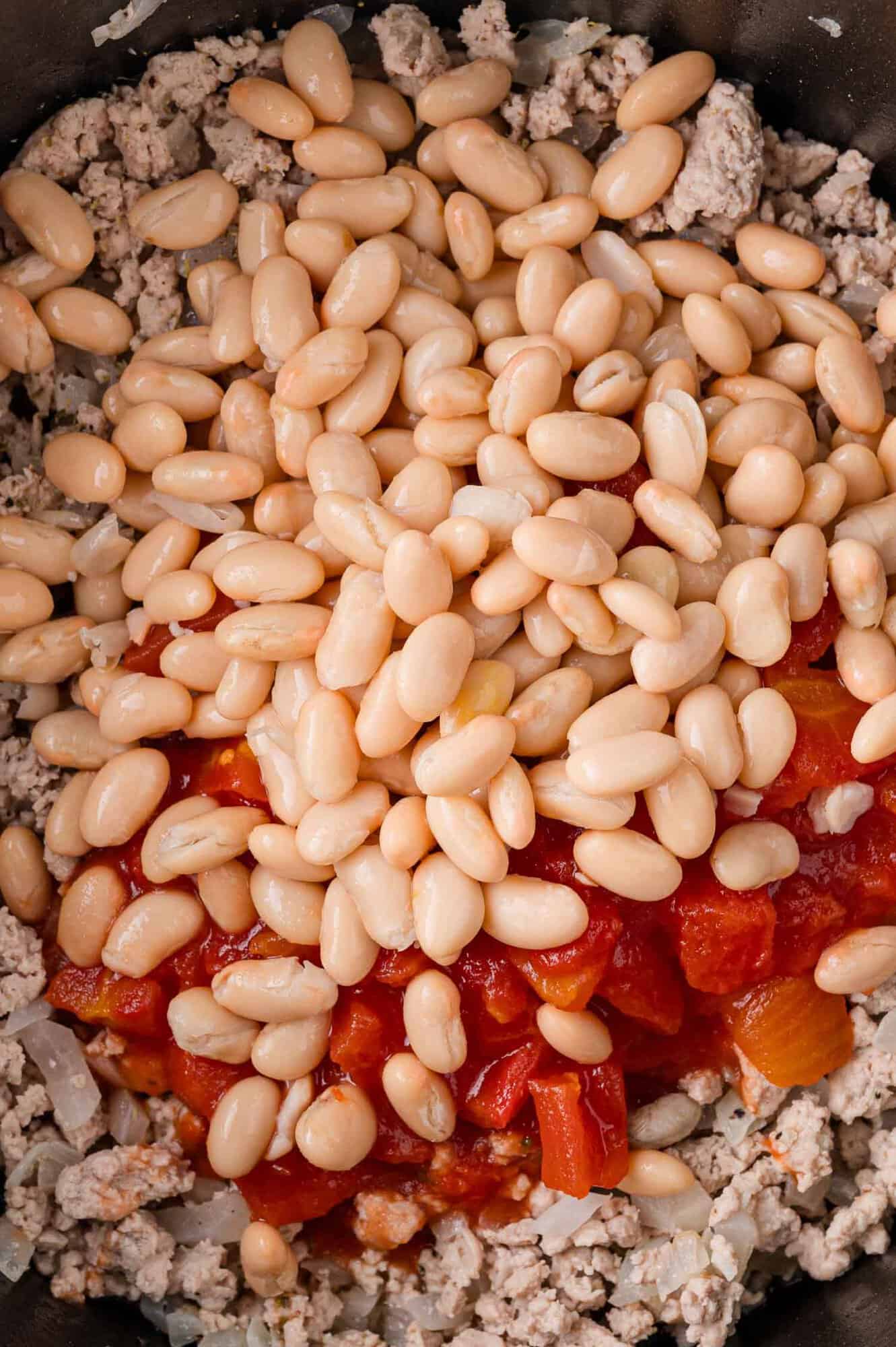Close-up of white beans and diced tomatoes on top of cooked ground meat and onions.