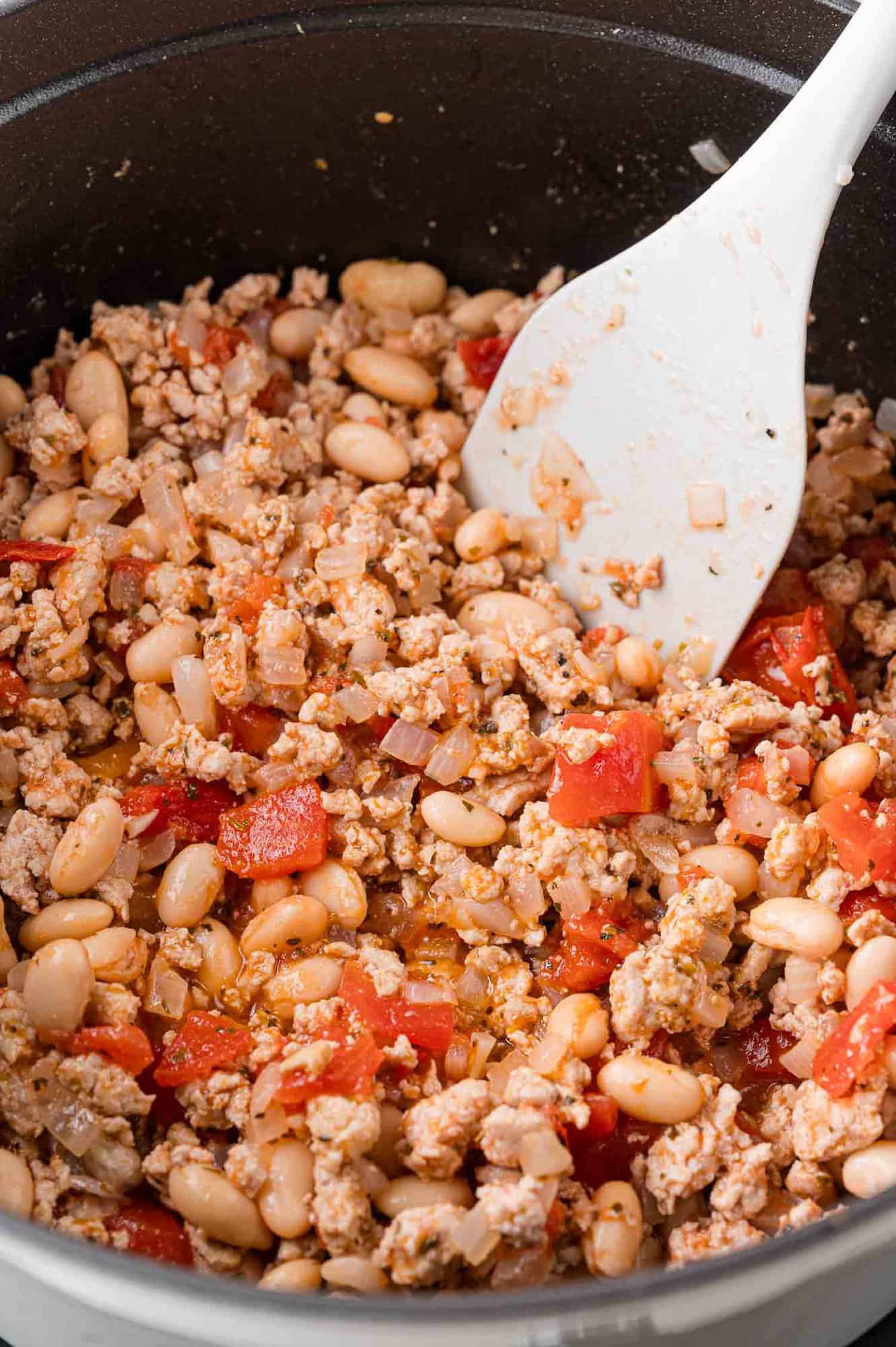 A pot containing cooked ground meat, white beans, diced tomatoes, and onions, with a white spatula resting inside.