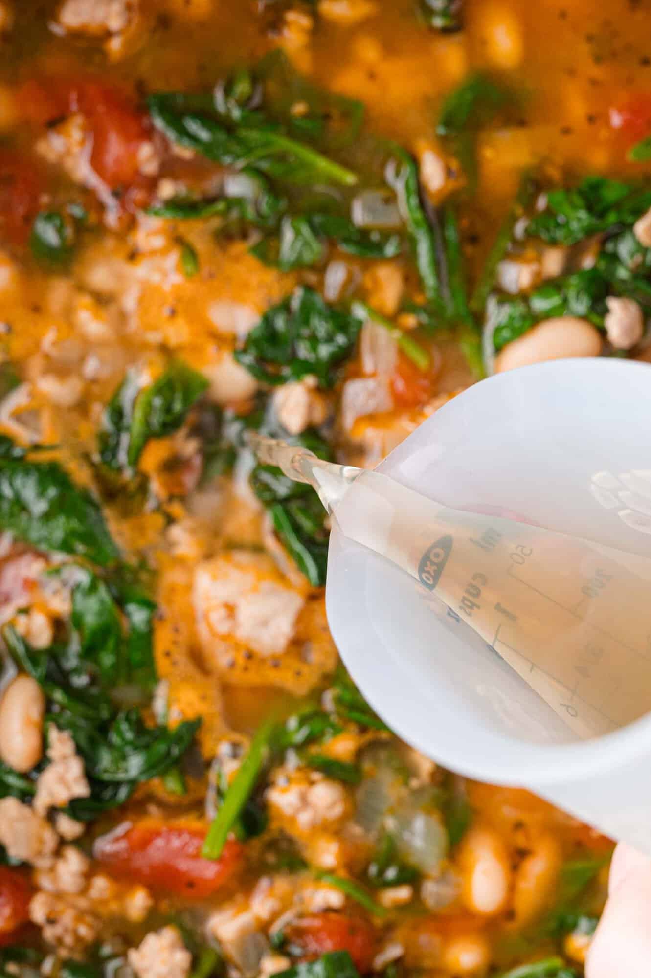 Pouring broth into a pot with spinach, tomatoes, beans, and ground meat.