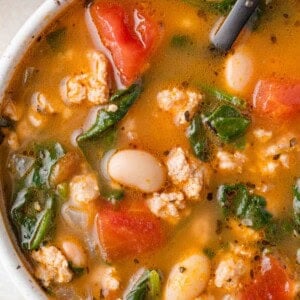 A bowl of tomato-based soup with white beans, greens, and ground meat, served with a black spoon.