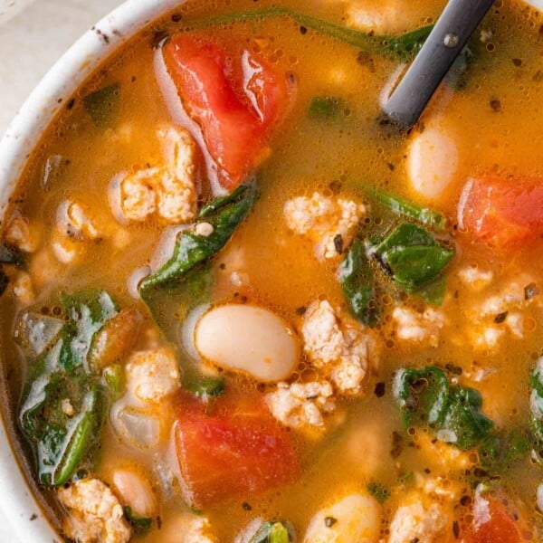 Bowl of soup with chunks of tomato, white beans, spinach, and ground meat in a broth, with a spoon.