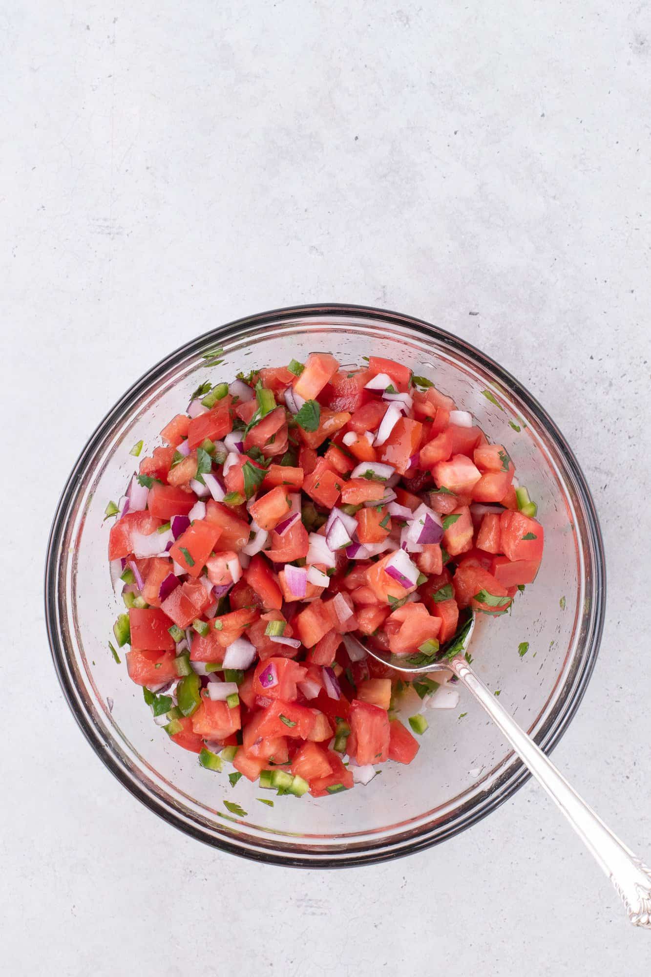 A clear bowl filled with fresh diced tomatoes, onions, jalapeños, cilantro, and a spoon on a white surface.