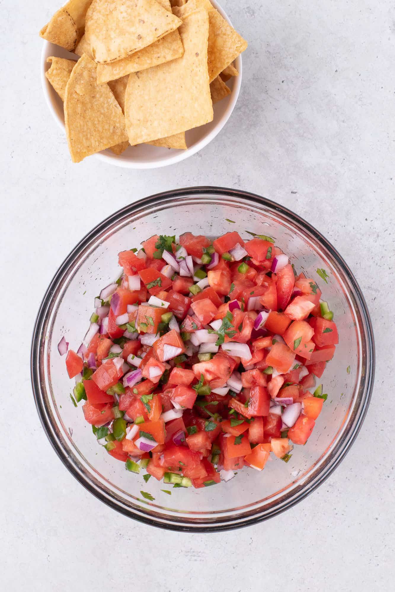 A bowl of pico de gallo with diced tomatoes, onions, and cilantro, next to a bowl of tortilla chips on a light surface.