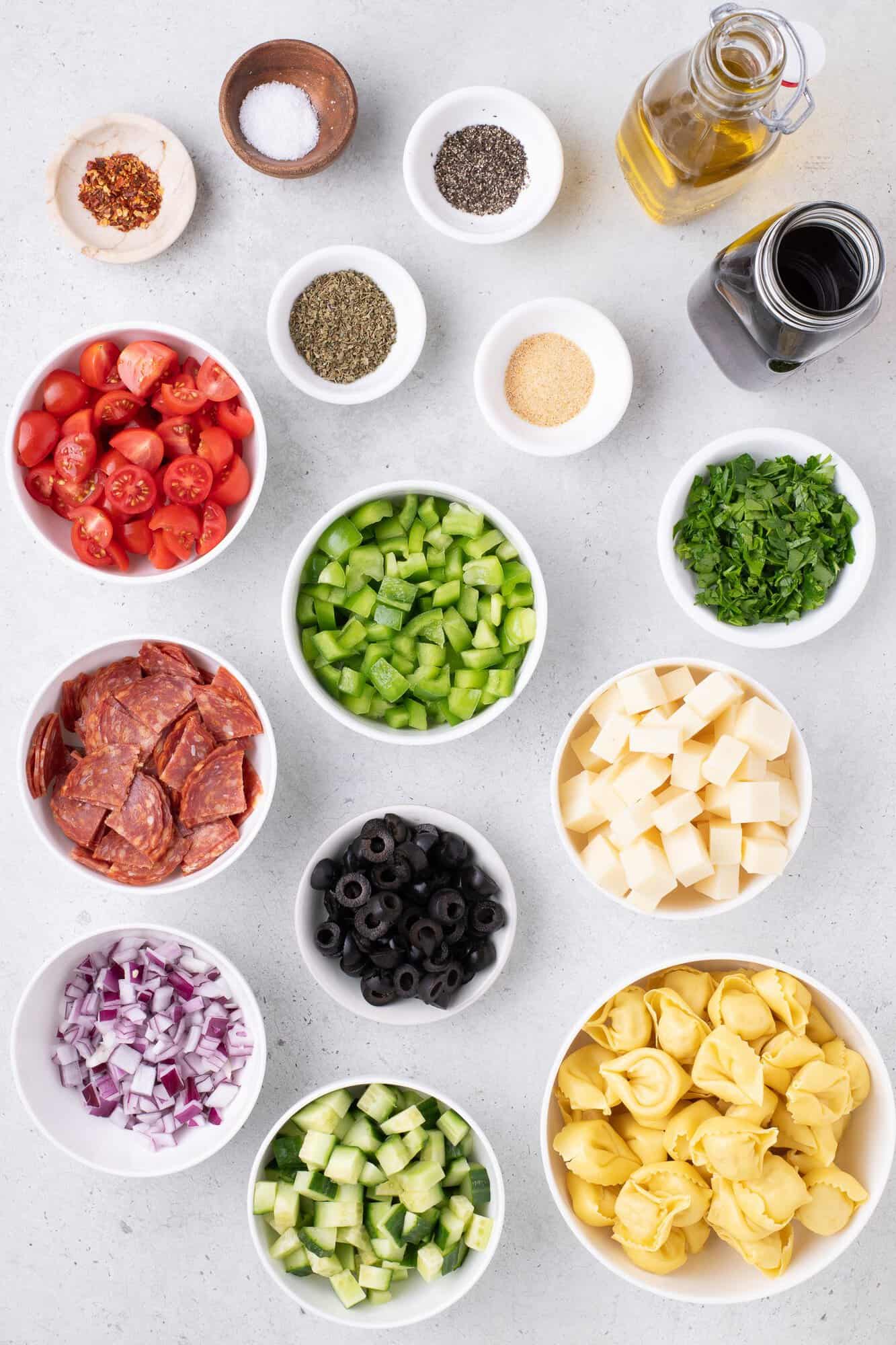 Variety of ingredients on a table, including tortellini, cheese cubes, black olives, green peppers, cherry tomatoes, cucumber, red onion, pepperoni, and seasonings.