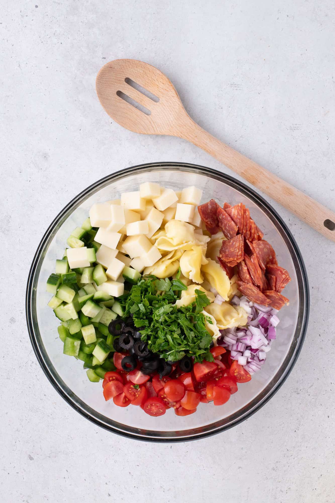 A glass bowl with chopped cucumbers, mozzarella, tortellini, pepperoni, black olives, tomatoes, red onion, and parsley. A wooden spoon lies beside the bowl.