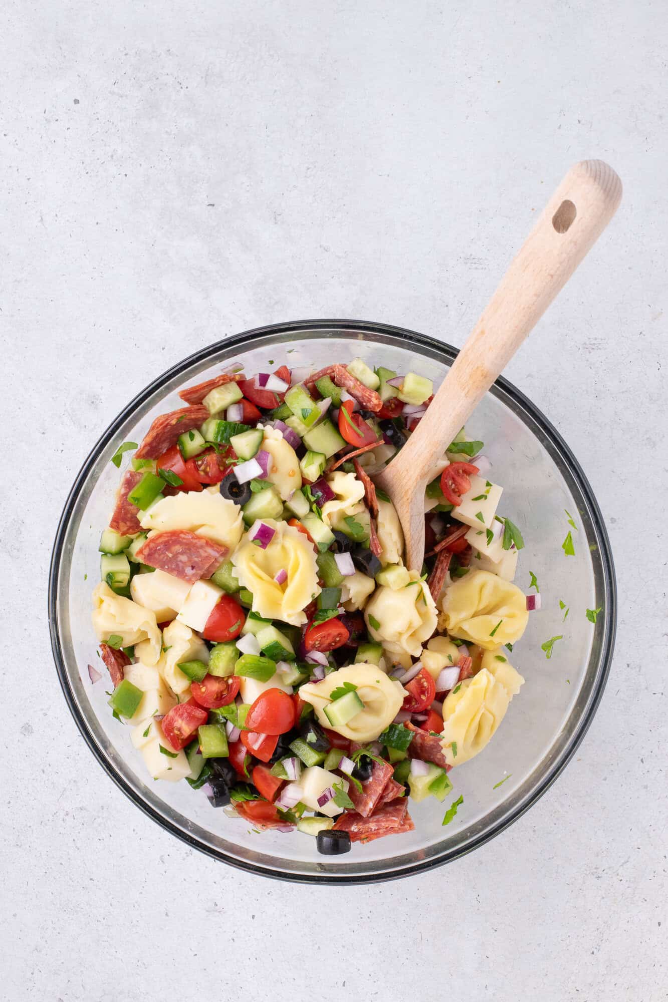 A bowl of tortellini salad with chopped vegetables, salami, and cheese, with a wooden spoon on a light-colored surface.