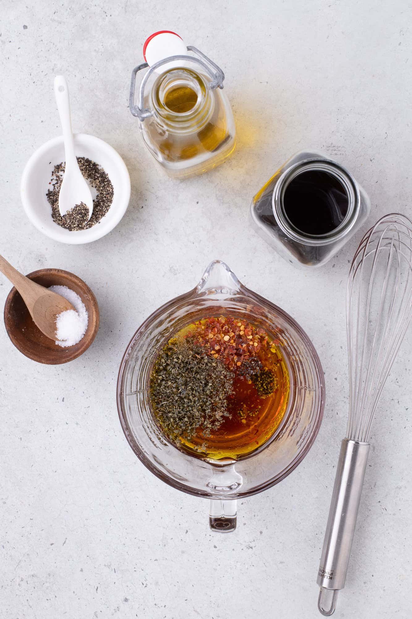 Ingredients for a dressing are placed on a light surface: a glass bowl with oil and spices, a whisk, and small bowls with salt, pepper, and herbs. Two jars of oil and vinegar are nearby.