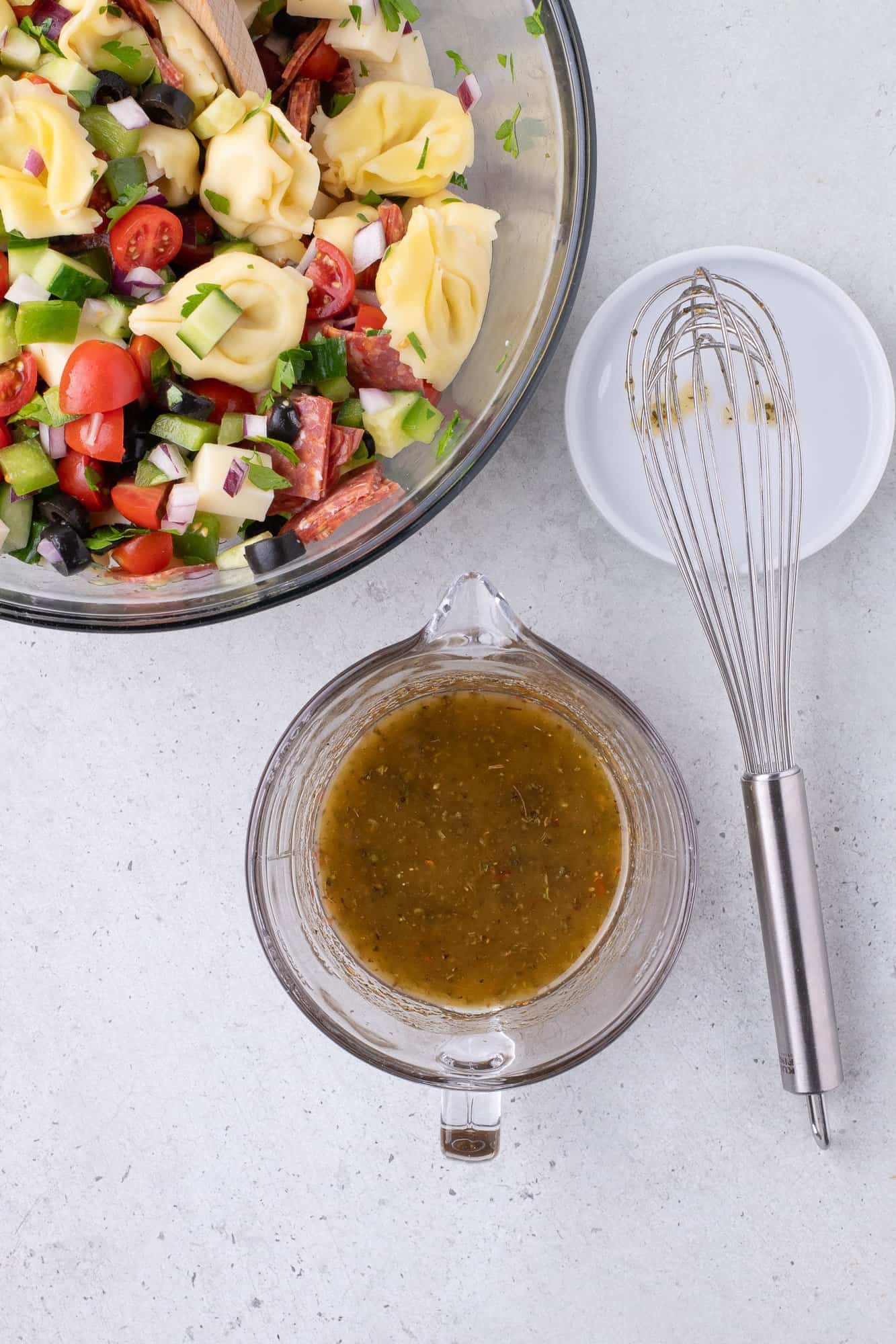 A bowl of tortellini salad with cherry tomatoes, cucumbers, bell peppers, and olives next to a whisk and bowl of dressing.
