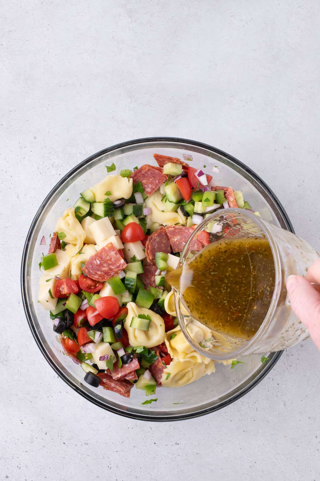 A hand pours dressing over a salad with tortellini, salami, tomatoes, cheese, cucumbers, olives, and onions in a glass bowl.