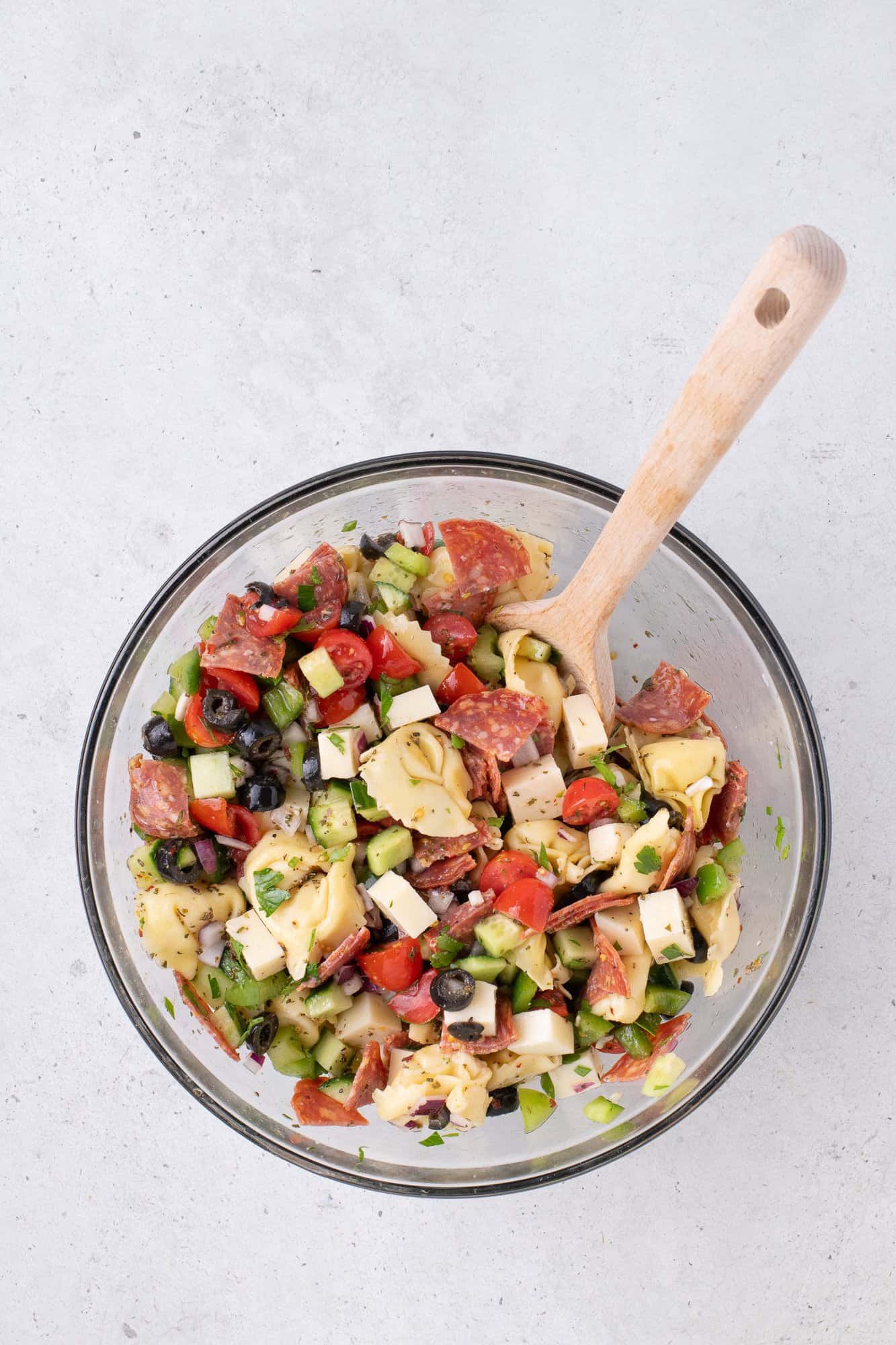 A glass bowl filled with a chopped salad containing tomatoes, cucumbers, olives, cheese, and meat slices, with a wooden spoon.