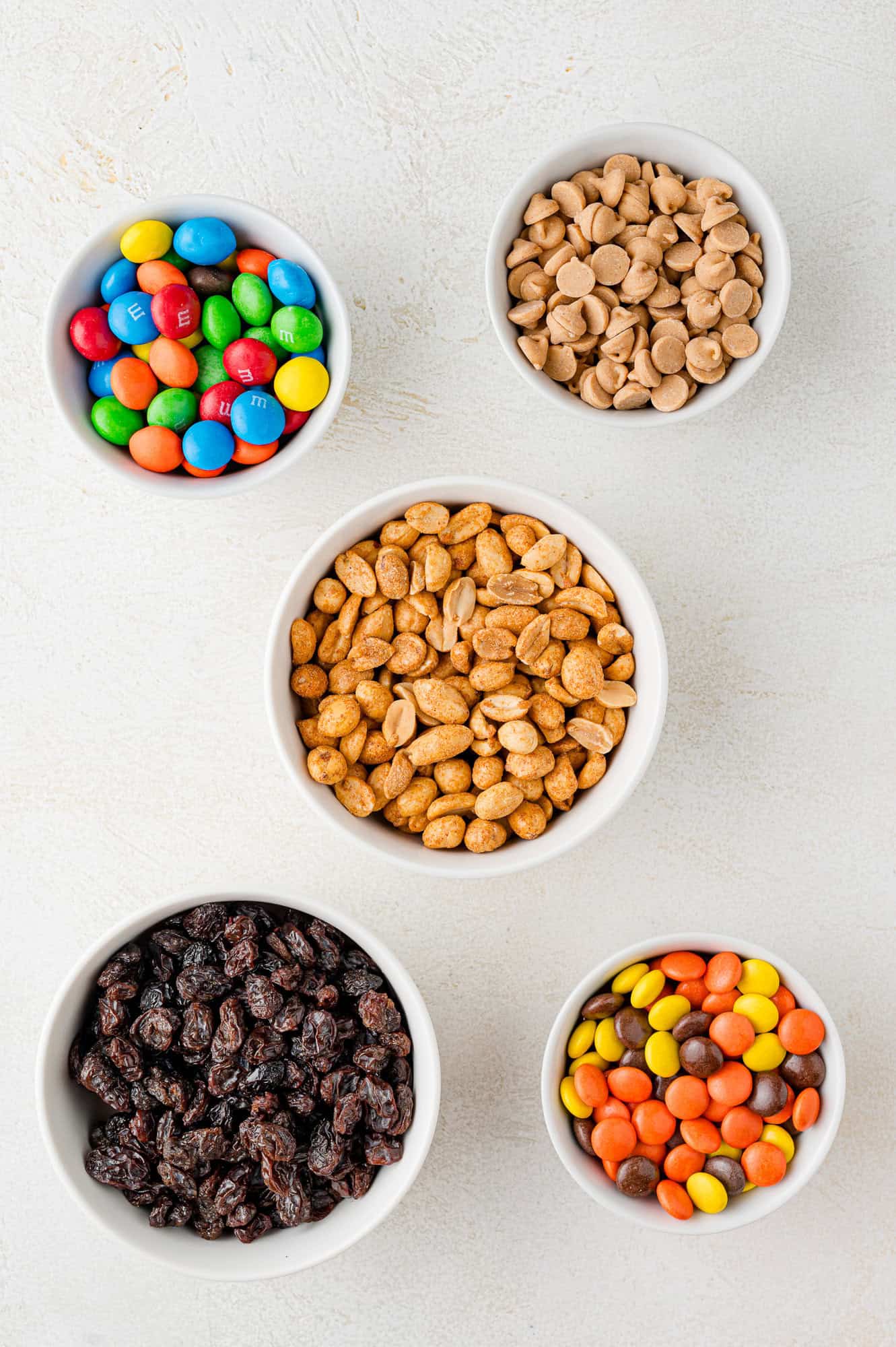 Five bowls with colorful candies, peanut butter chips, peanuts, chocolate candies, and raisins arranged on a light surface.