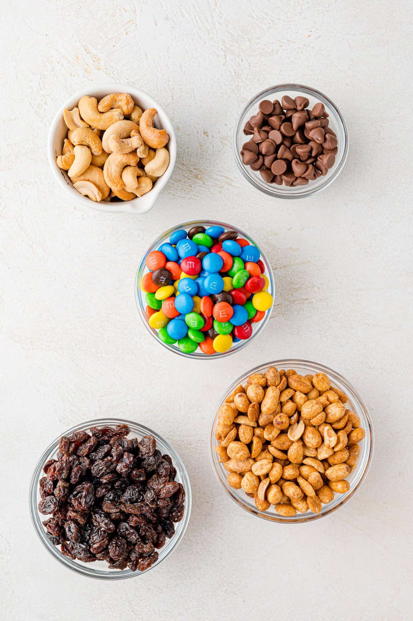 Five bowls containing cashews, chocolate chips, M&Ms, raisins, and peanuts arranged on a white surface.