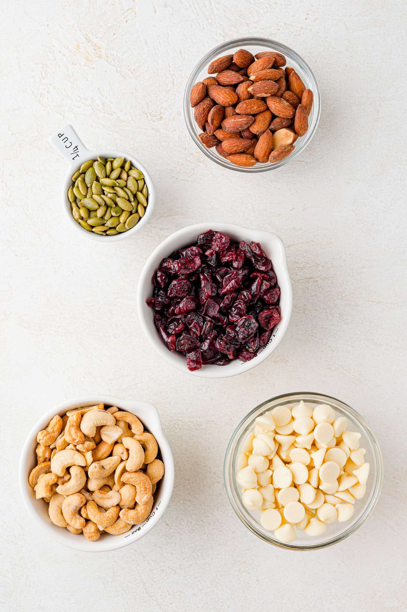 Bowls of almonds, cashews, dried cranberries, white chocolate chips, and pumpkin seeds on a white surface.