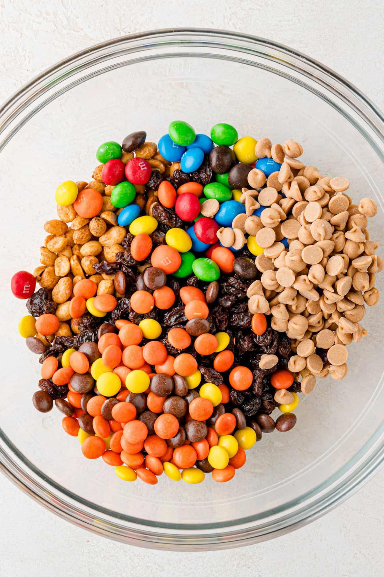 A glass bowl filled with colorful candy-coated chocolate, peanuts, raisins, and peanut butter chips.