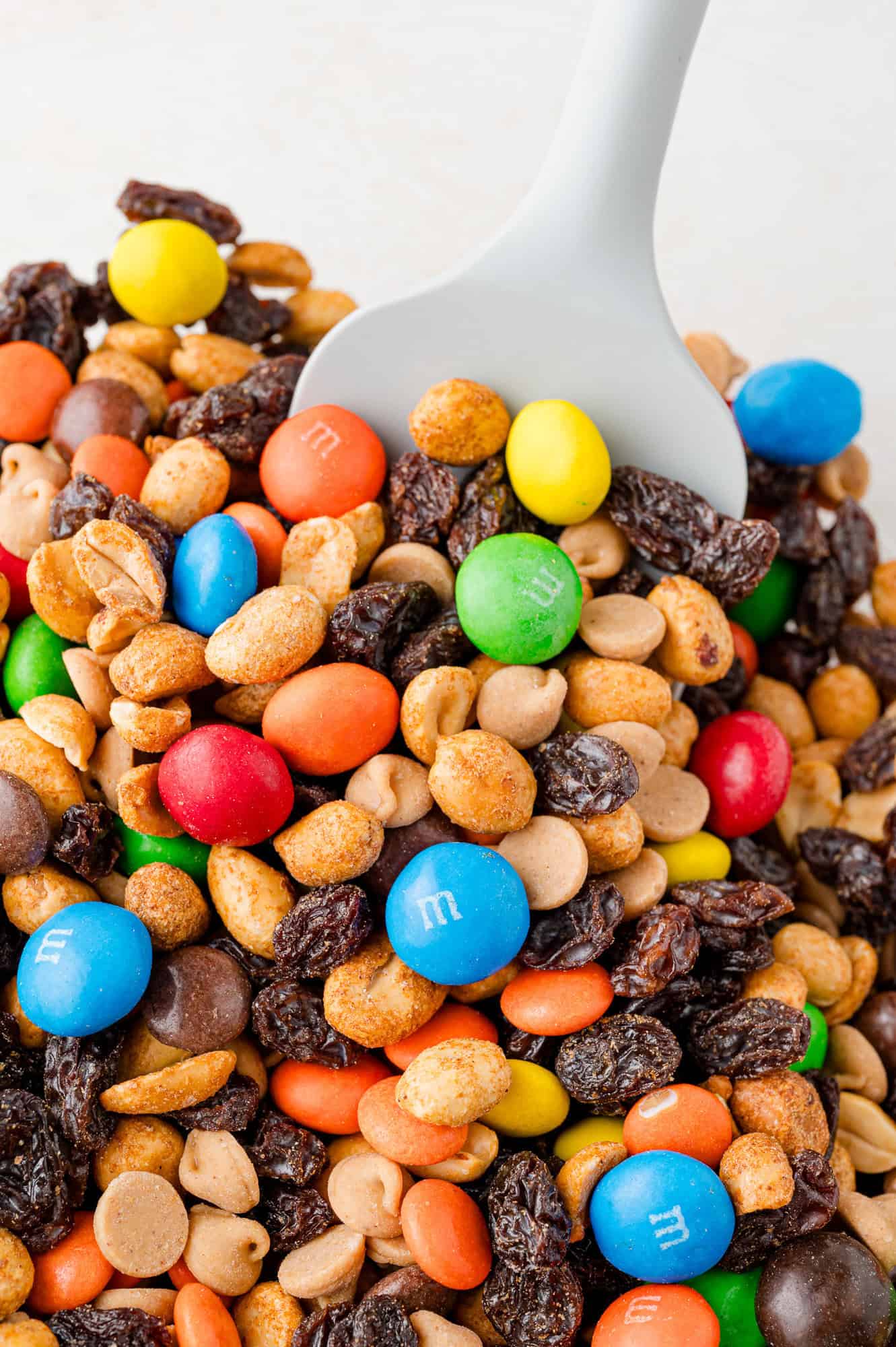 A scoop of trail mix with raisins, colored candy-coated chocolates, peanuts, and other assorted treats.