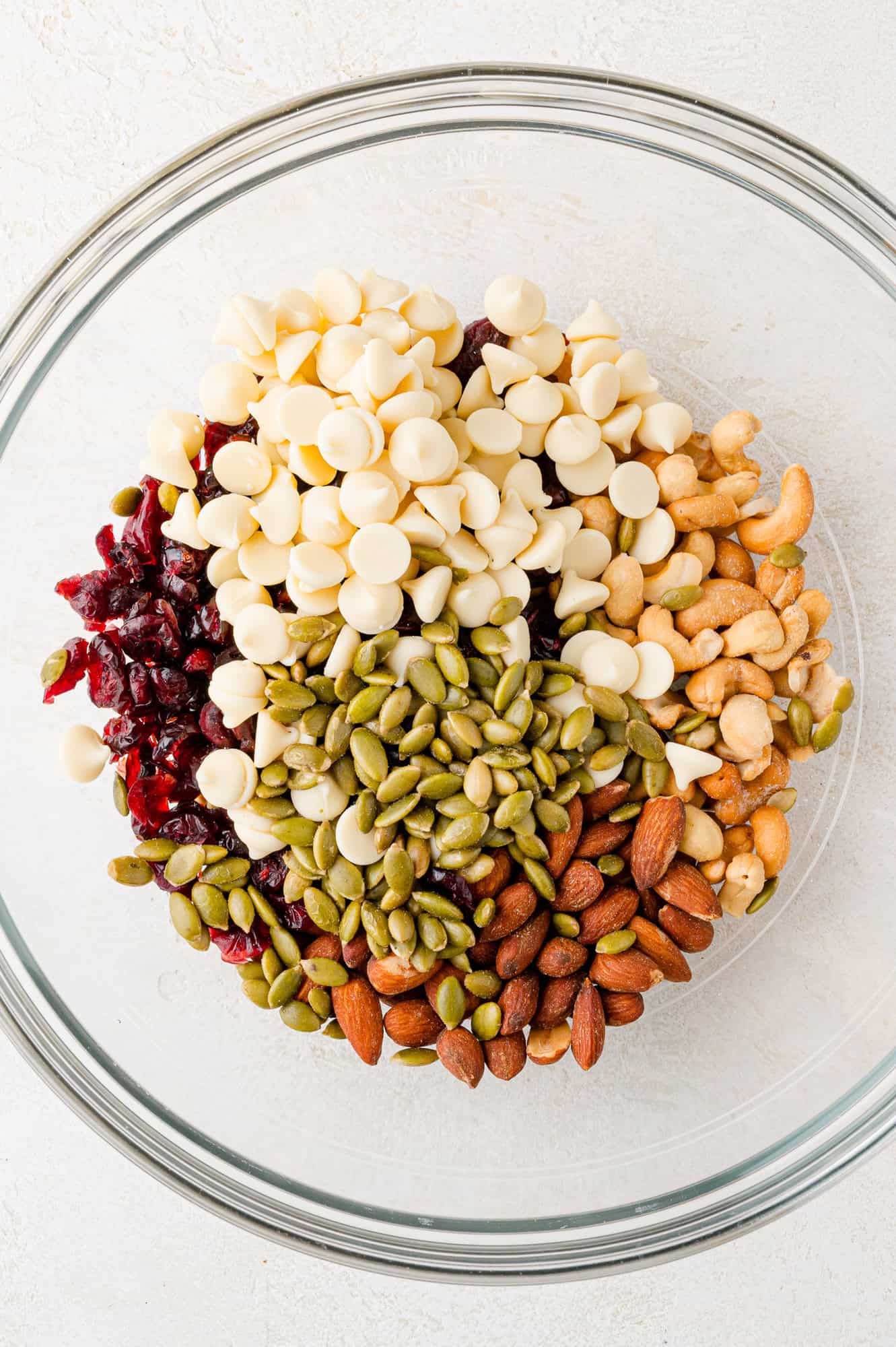 A glass bowl containing white chocolate chips, dried cranberries, pumpkin seeds, almonds, and cashews on a light surface.
