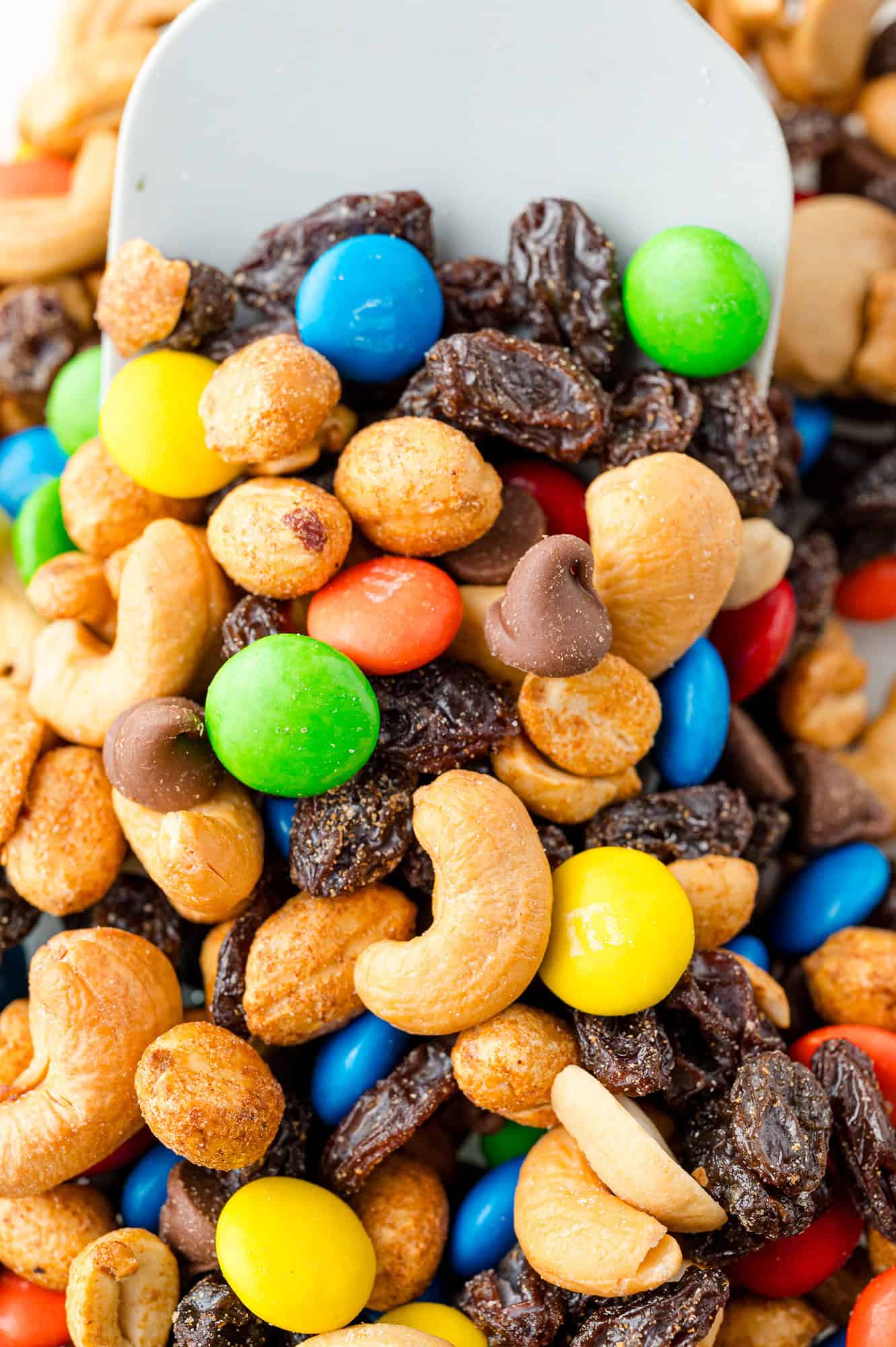 A close-up of trail mix with cashews, raisins, chocolate candies, and peanuts, mixed together with a light-colored scoop.