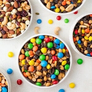 Bowls of assorted trail mix with nuts, candies, and raisins on a white surface.
