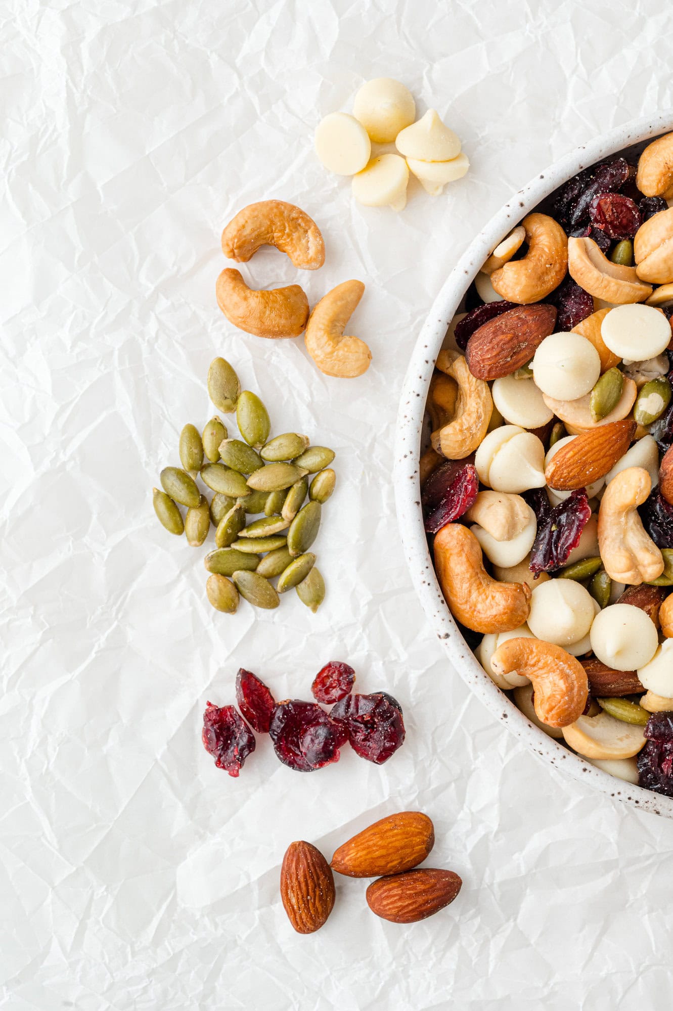 A bowl of mixed nuts with dried cranberries on a white surface, surrounded by clusters of cashews, pumpkin seeds, white chocolate chips, almonds, and dried cranberries.
