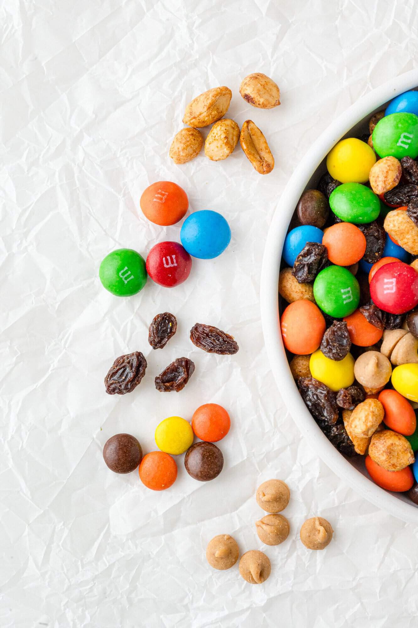 A bowl of M&Ms, nuts, and raisins on crinkled white paper, with some scattered outside the bowl.