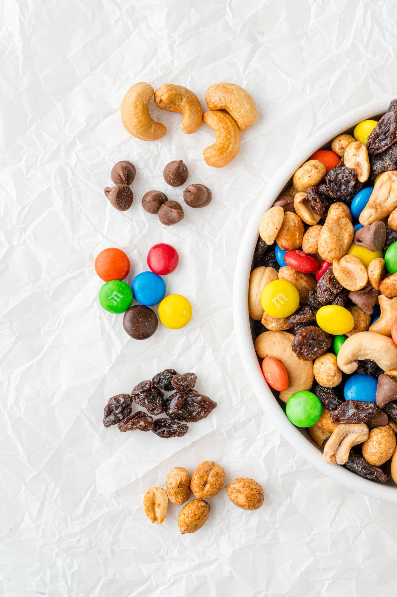 A white bowl of trail mix with assorted nuts, chocolate candies, and raisins, arranged in neat rows beside the bowl on a crinkled white surface.