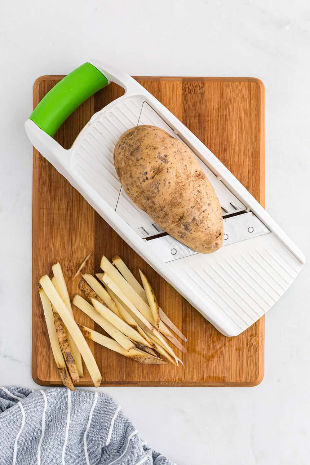 A potato on a mandoline slicer with cut potato strips beside it, placed on a wooden cutting board. A striped cloth is partially visible in the lower left corner.