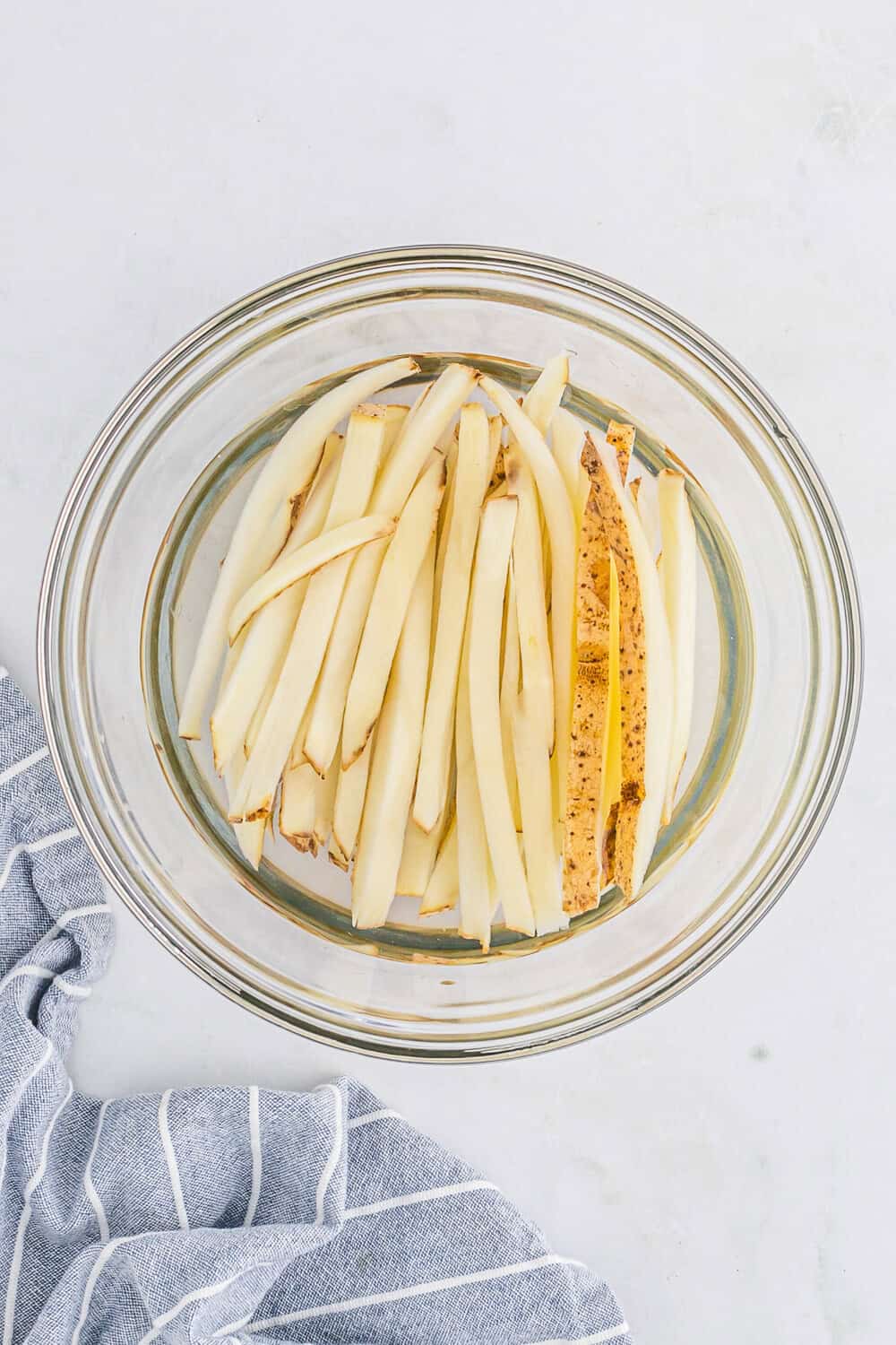 Potato slices soaking in water in a clear glass bowl, next to a striped cloth on a pale surface.