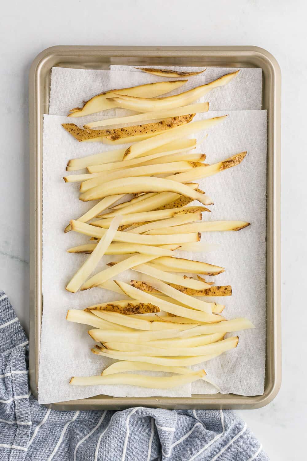Raw potato strips on a baking sheet lined with parchment paper, atop a striped cloth.