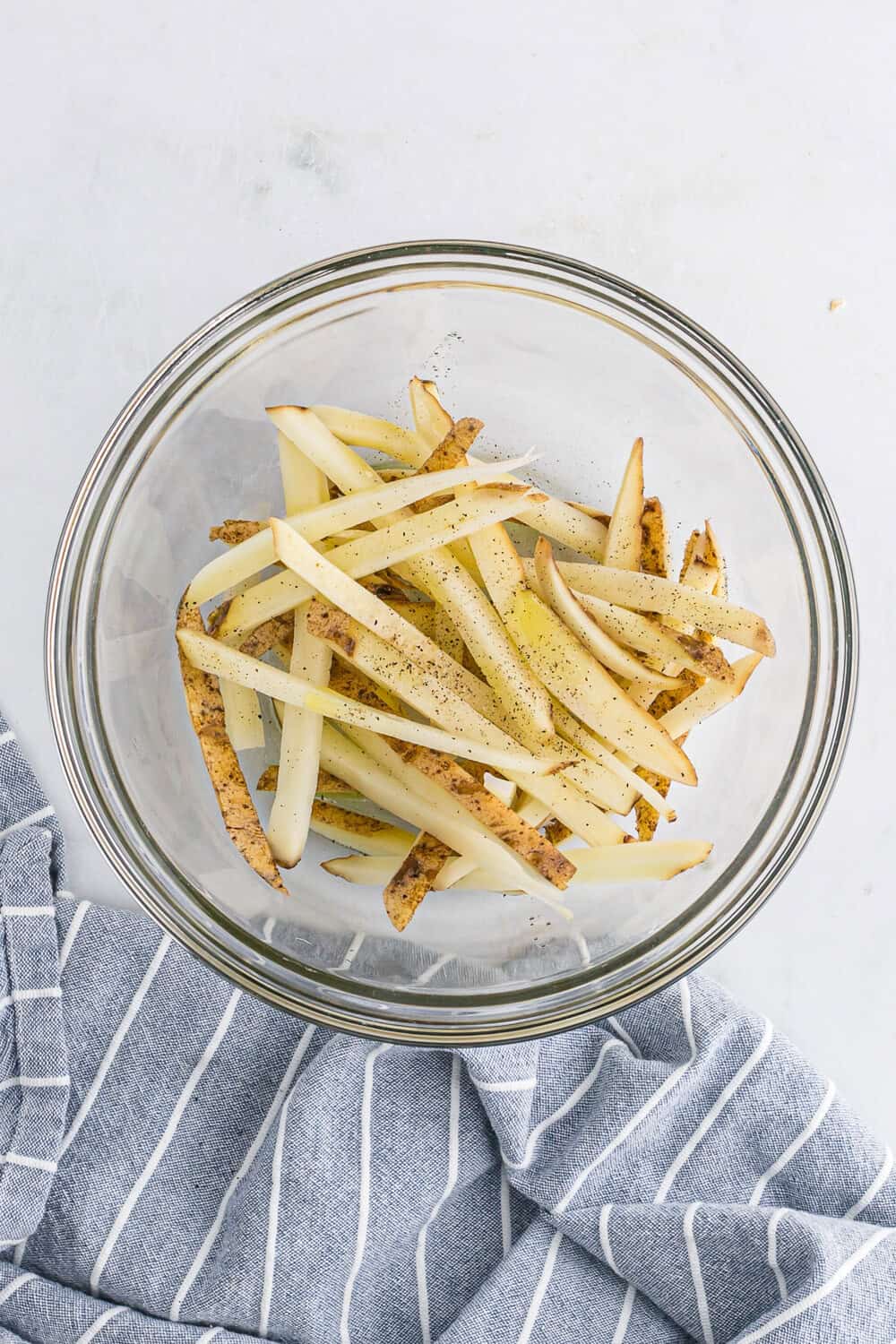 Potato slices seasoned with salt and pepper in a glass bowl on a gray striped cloth.