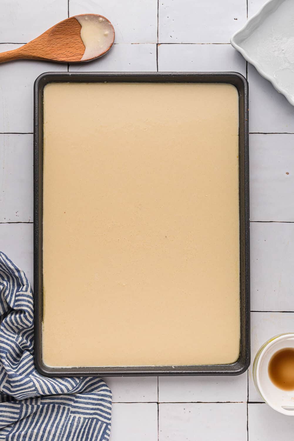 A baking sheet filled with liquid cake batter, surrounded by a wooden spoon, a striped cloth, a square baking dish, and a white cup on a tiled surface.