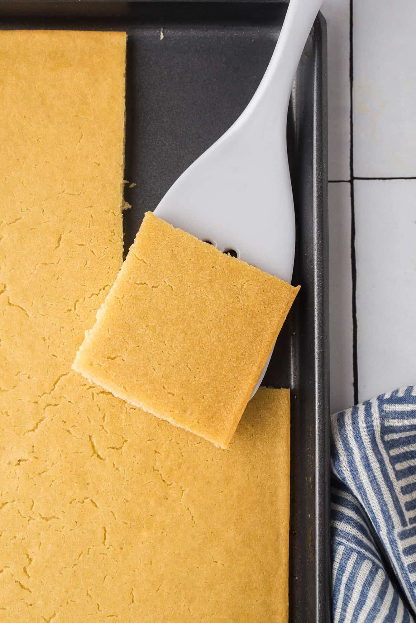 A baking tray with a square piece of pancake lifted by a white spatula. A striped cloth is partially visible in the corner.