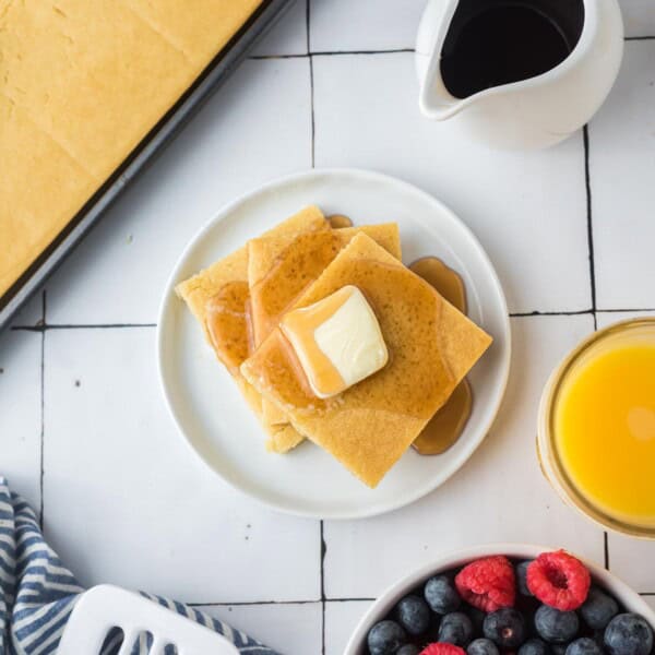 Two square pancakes topped with butter and syrup on a plate, next to a pitcher of syrup, a glass of orange juice, and a bowl of berries on a tiled surface.