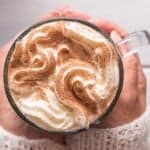 A person holding a glass mug of whipped cream-topped cinnamon latte, sprinkled with cinnamon.