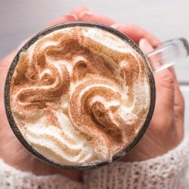 A person holding a glass mug of whipped cream-topped cinnamon latte, sprinkled with cinnamon.