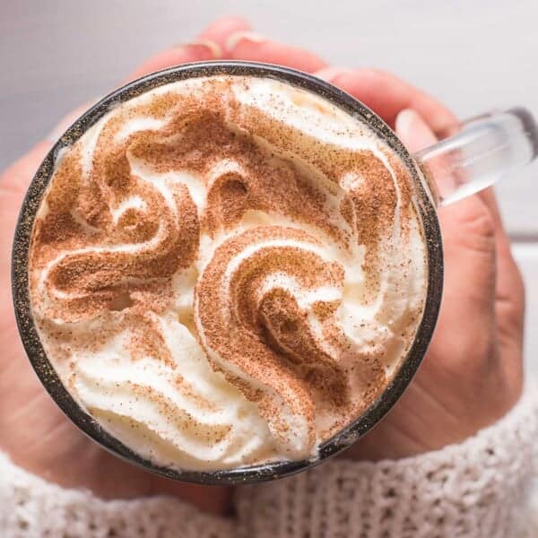 A person holding a glass mug of whipped cream-topped cinnamon latte, sprinkled with cinnamon.