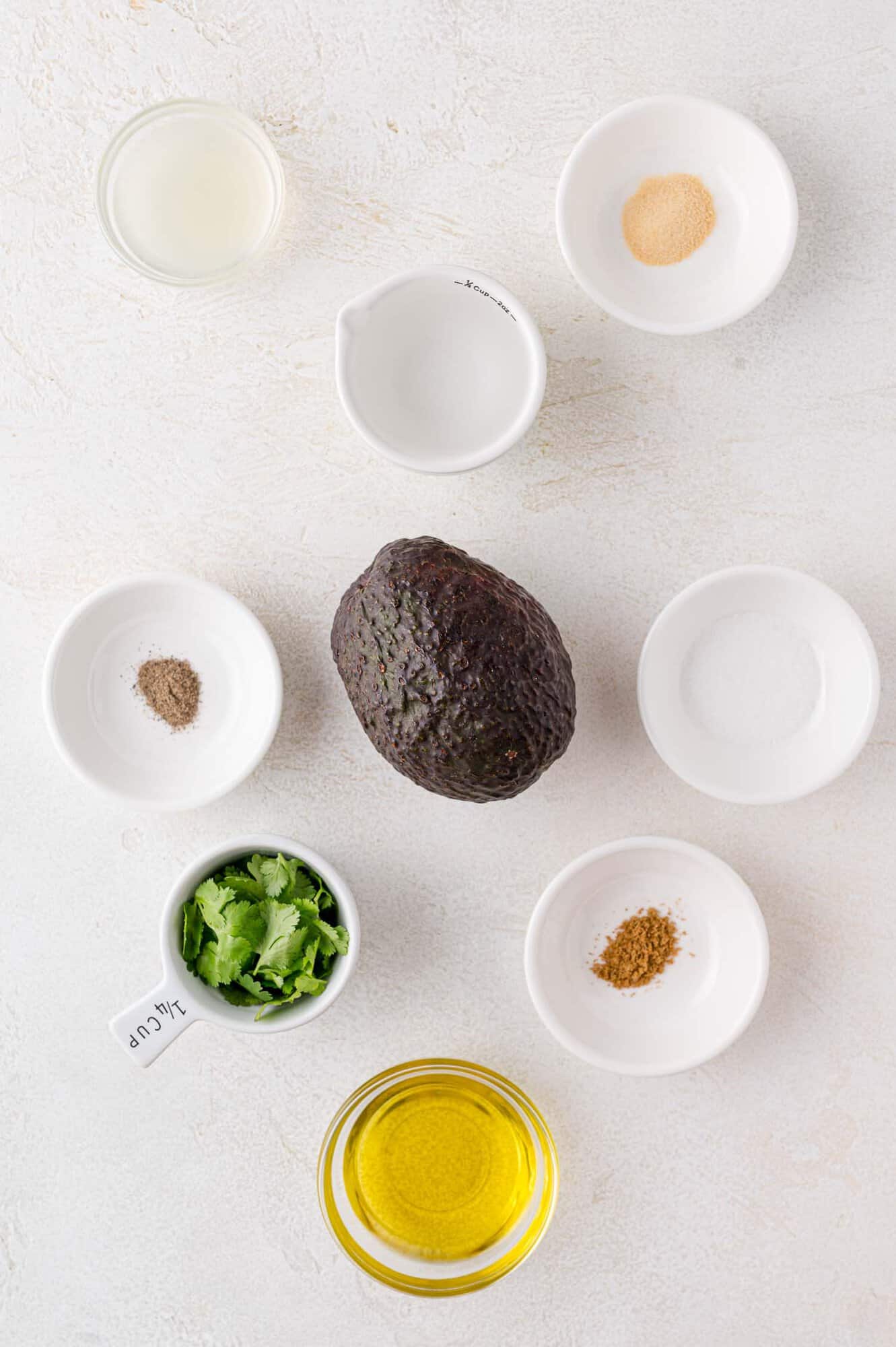 Ingredients for avocado dressing arranged on a surface: a whole avocado, olive oil, cilantro, lime juice, and seasonings in small bowls.