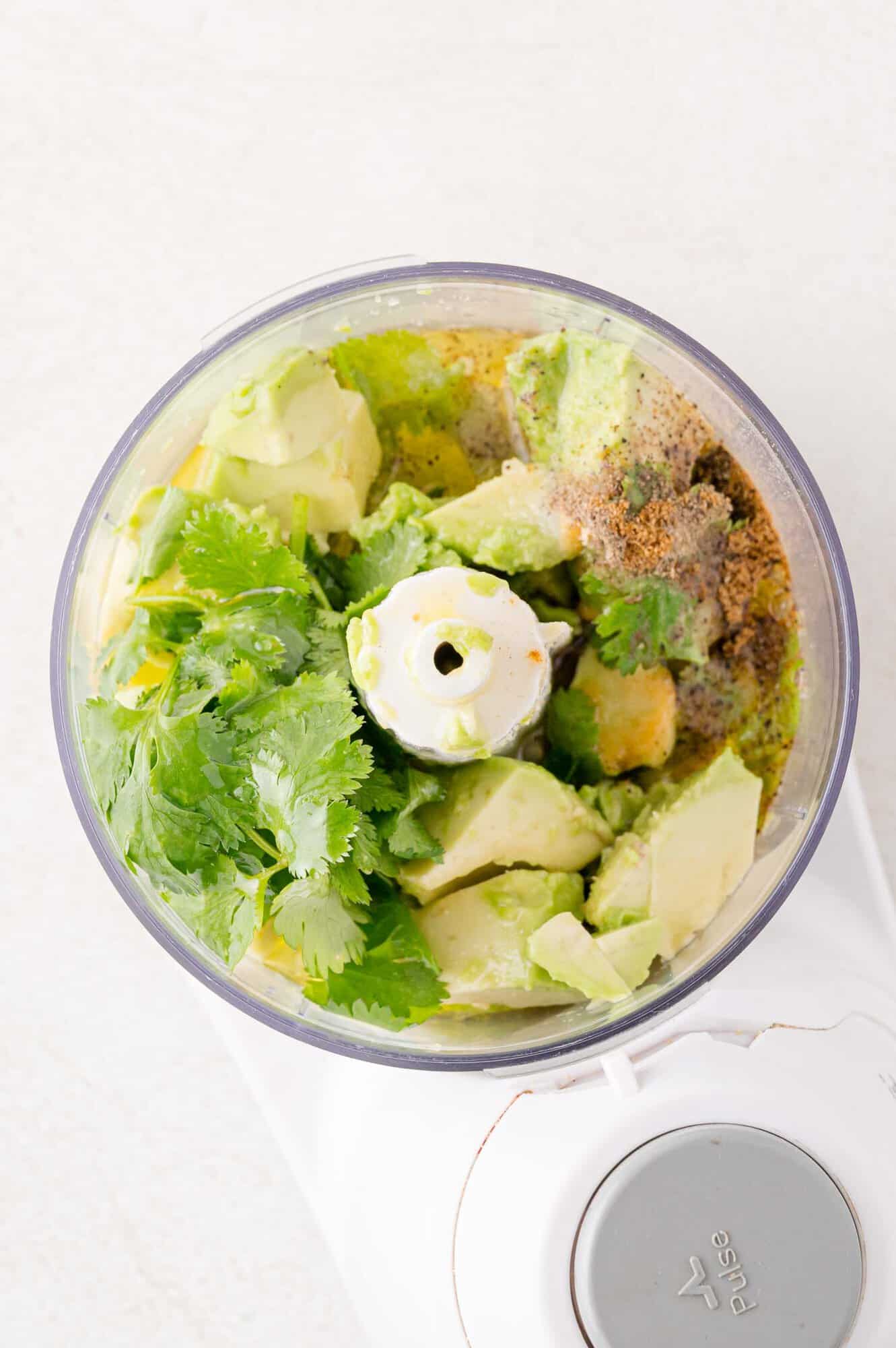 Food processor with avocado, cilantro, and spices inside, ready to be blended.