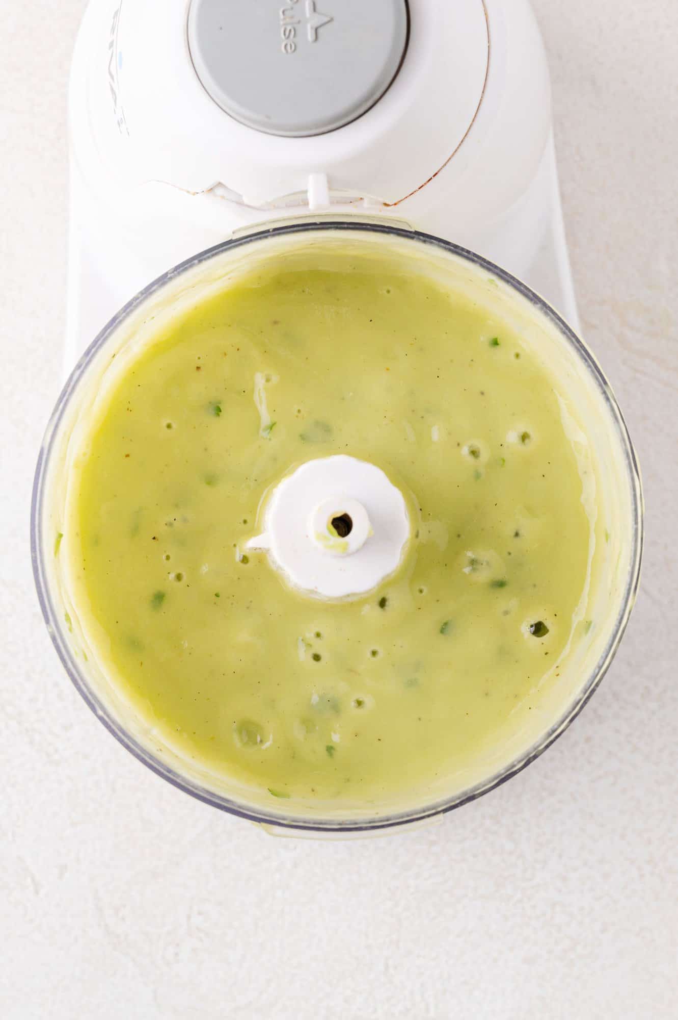 Top-down view of a food processor containing smooth green sauce with herbs, against a light background.