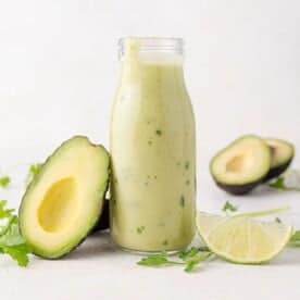 A bottle of green avocado dressing is placed next to an avocado half and a lime wedge, with cilantro garnish on a light surface.