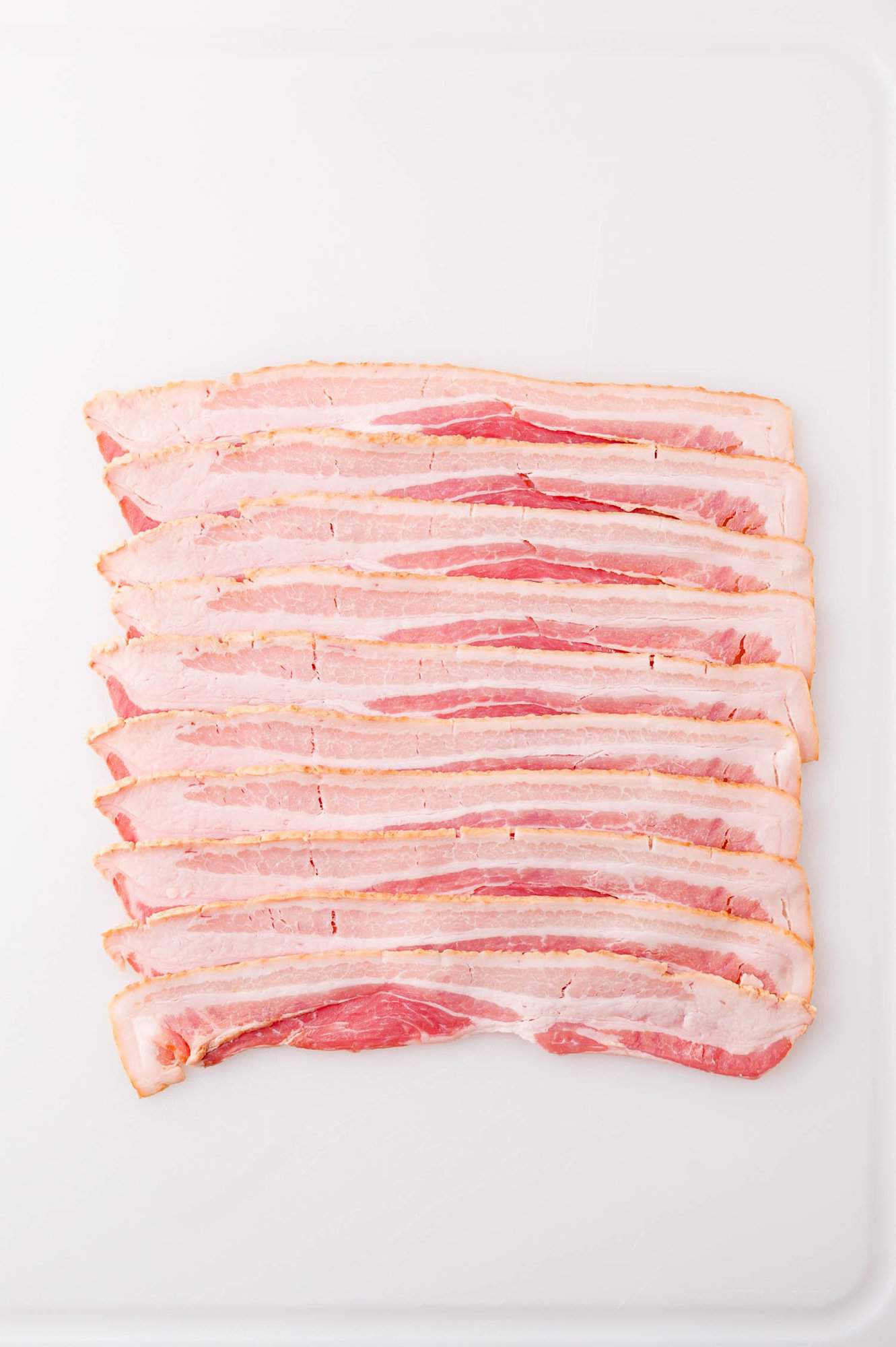 Several uncooked bacon strips are laid out in a neat row on a white surface.