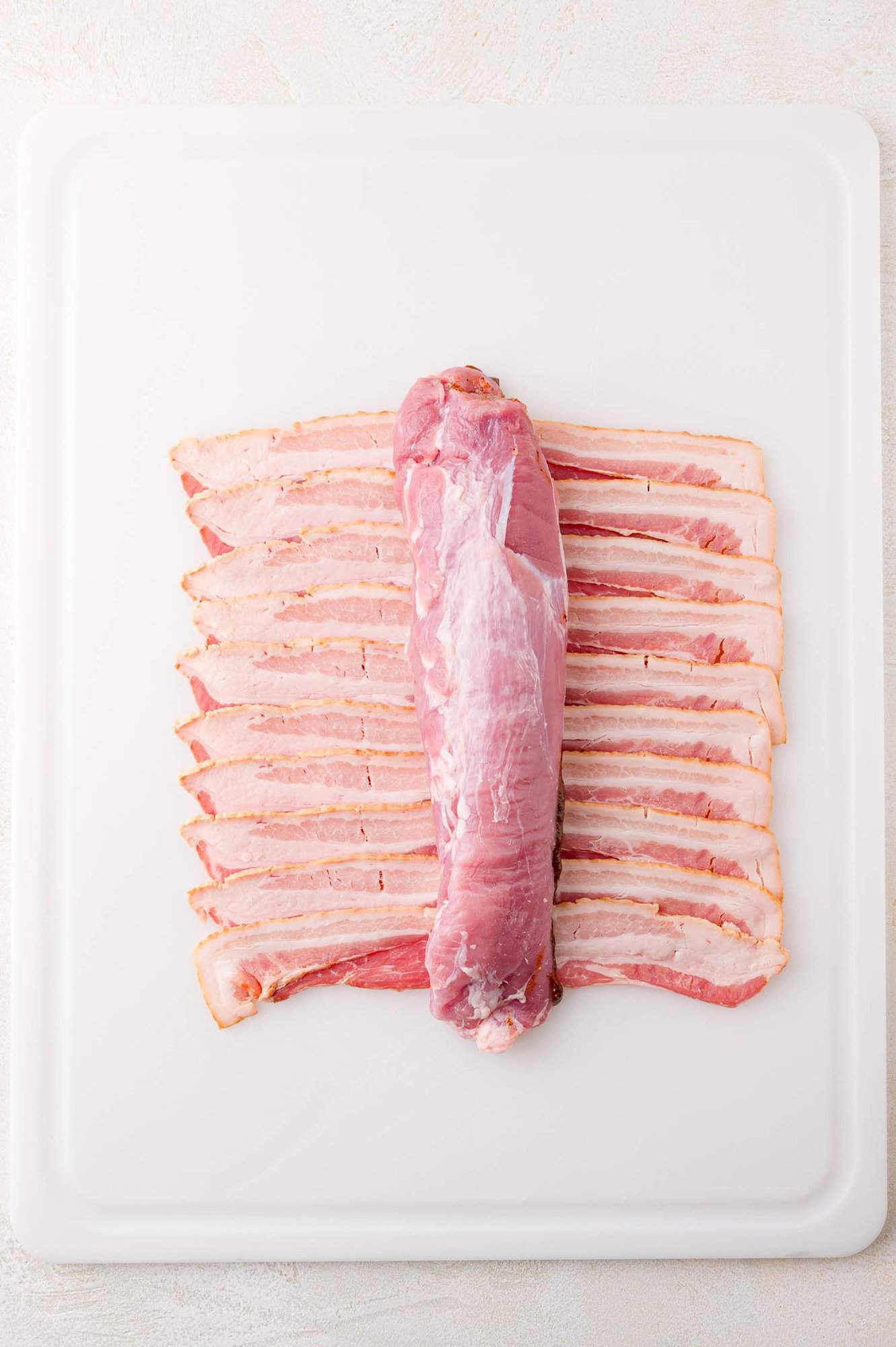 A raw pork tenderloin is placed on top of several strips of uncooked bacon, arranged neatly in rows, on a white cutting board.
