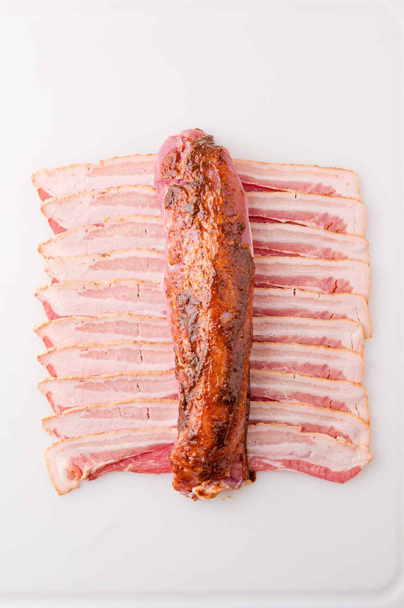 A row of sliced bacon is laid out on a white surface with a seasoned pork tenderloin placed on top.
