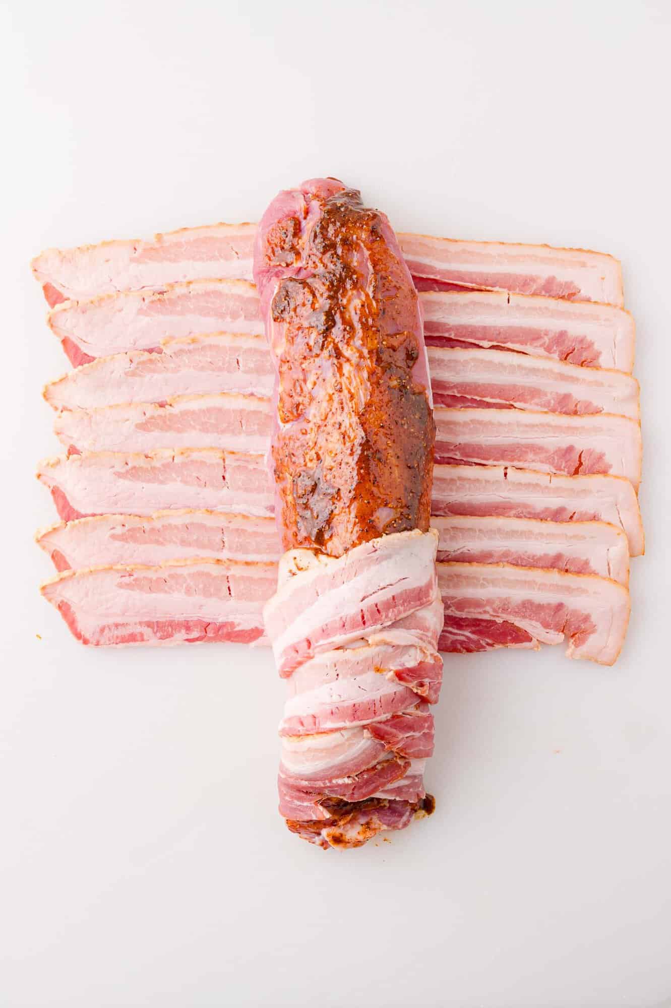 Raw pork tenderloin seasoned and wrapped with strips of uncooked bacon, displayed on a white surface.