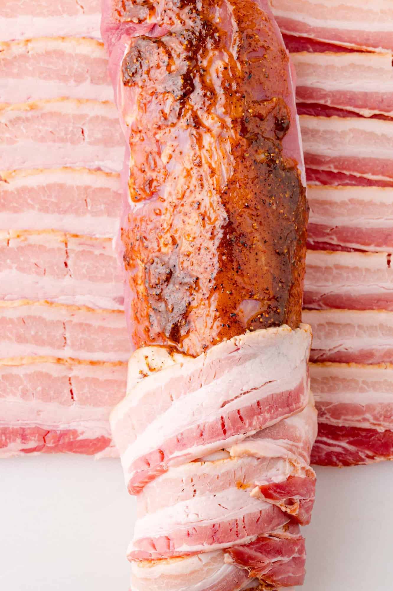 Raw pork tenderloin wrapped in bacon slices, placed on a bed of additional bacon strips.