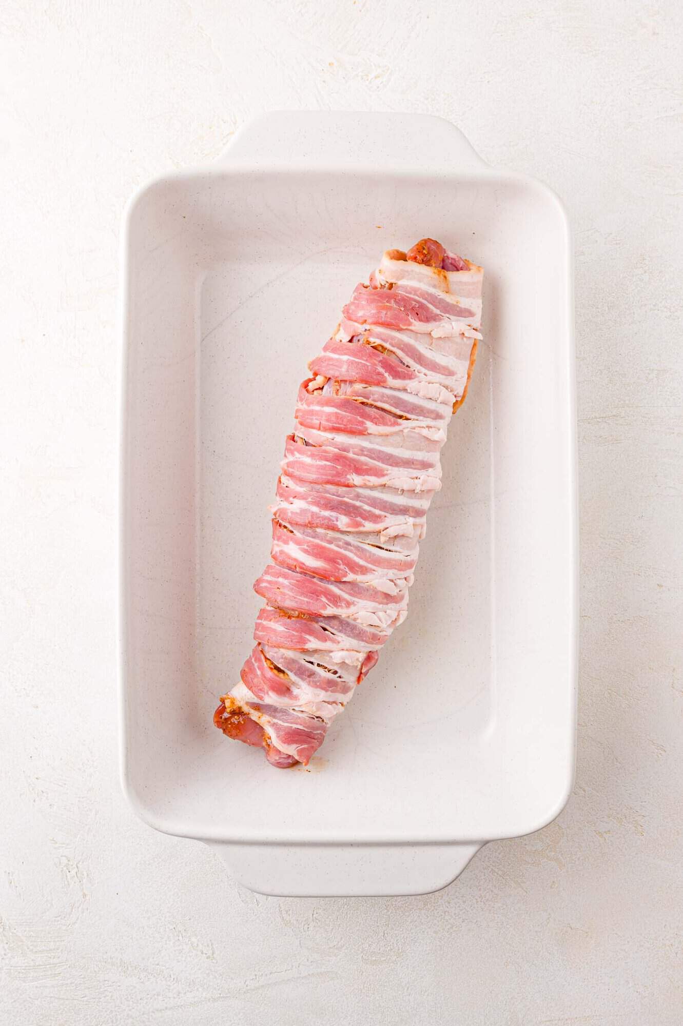 A piece of bacon-wrapped pork tenderloin on a white baking dish.
