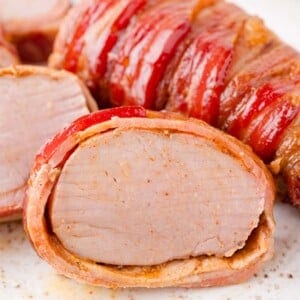 Sliced bacon-wrapped pork tenderloin on a white plate, showing a juicy interior and crispy bacon exterior.