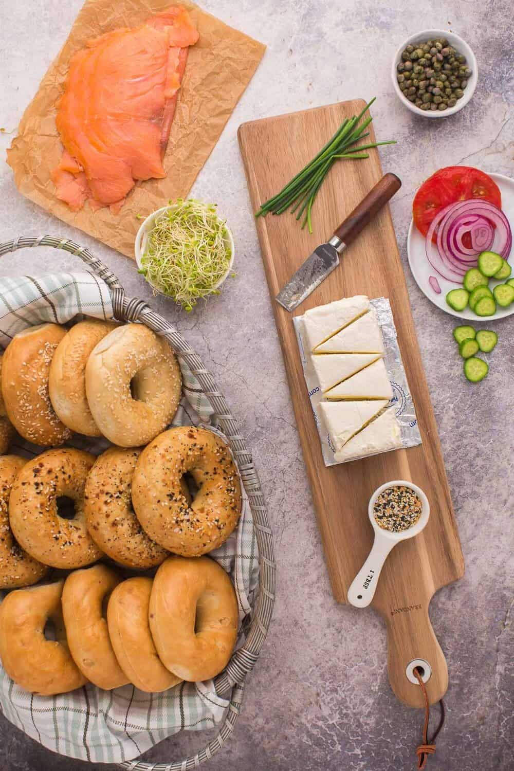Bagels in a basket next to smoked salmon, cream cheese, sliced tomatoes, red onions, cucumbers, sprouts, capers, and a knife with chives on a wooden board. Everything bagel seasoning is nearby.